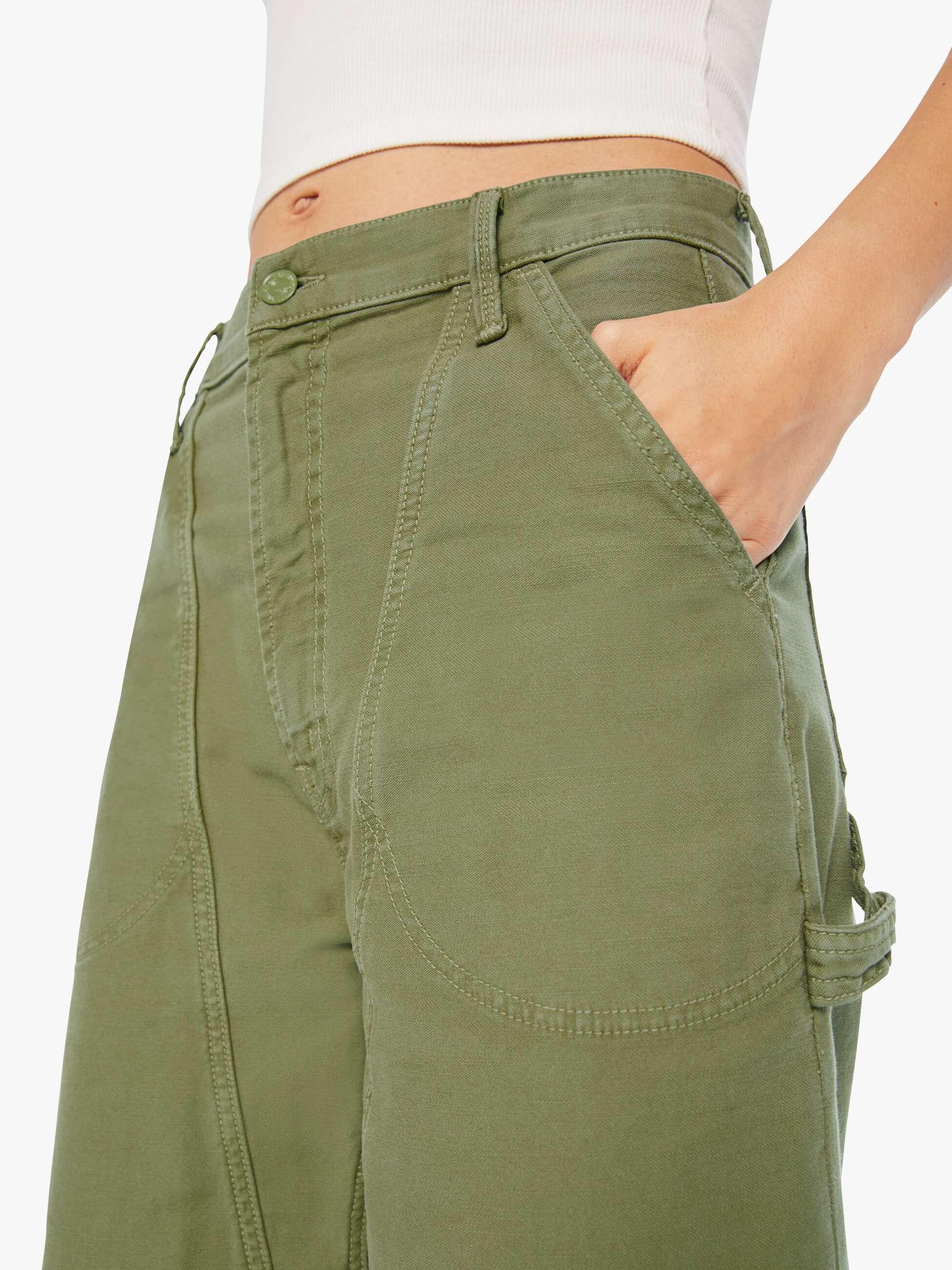 A person wears The Half Chisel high-waisted cargo pants by Mother in army green, featuring utility patch pockets and detailed stitching, paired with a white ribbed crop top. One hand rests in the pocket, highlighting the pants’ design.