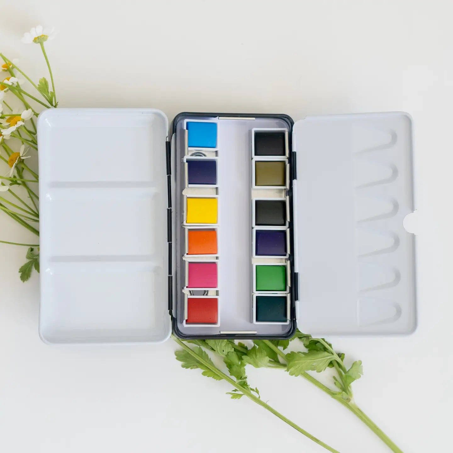 The Faire Travel Watercolor Paint Palette, featuring twelve vibrant pans, sits open on a white surface surrounded by art supplies, green leaves, and yellow-and-white flowers.