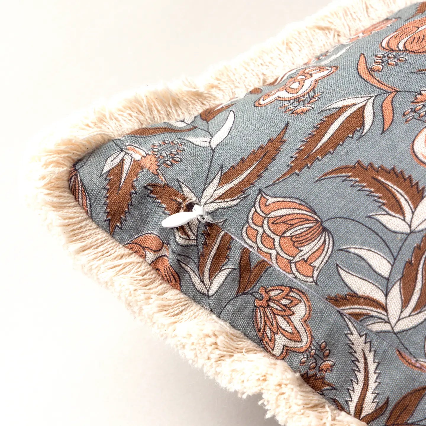 Close-up of the Block Print Pillow by Faire, featuring a blue base with brown and white floral patterns, beige fringe trim along the edge, and a white button at one corner.