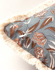 Close-up of the Block Print Pillow by Faire, featuring a blue base with brown and white floral patterns, beige fringe trim along the edge, and a white button at one corner.