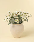A cream-colored vase displays Indaba’s Ranunculus Spray, White—realistic faux white blooms with green stems and leaves—set against a pale beige background.