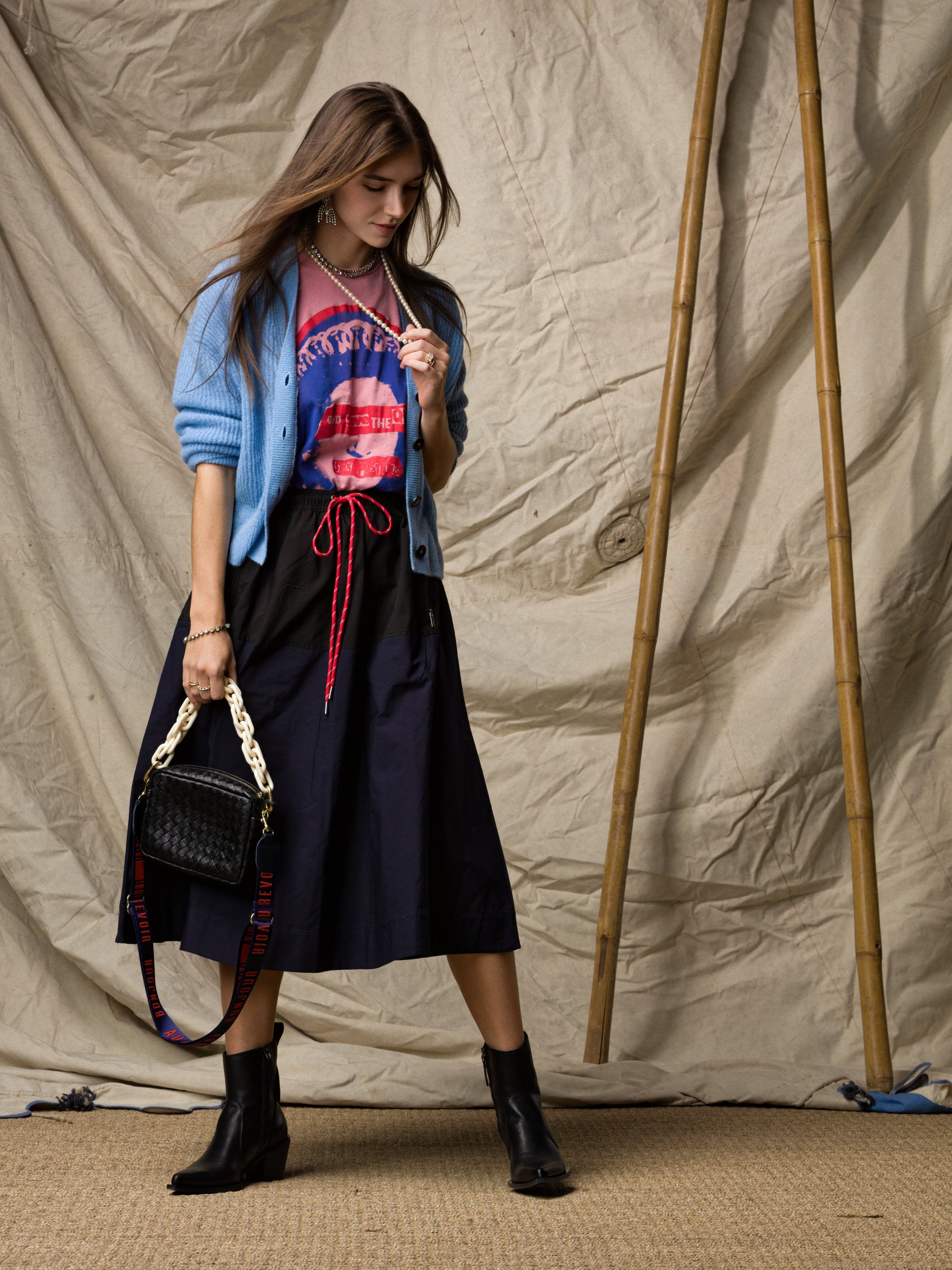 A woman stands before a beige backdrop in a light blue cardigan, Ping Pong Surf Club t-shirt, and the Le Vintage Sport Skirt by Ping Pong Surf Club with black ankle boots. She holds a black chain-strap handbag; bamboo poles accent the scene.