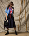 A woman stands before a beige backdrop in a light blue cardigan, Ping Pong Surf Club t-shirt, and the Le Vintage Sport Skirt by Ping Pong Surf Club with black ankle boots. She holds a black chain-strap handbag; bamboo poles accent the scene.