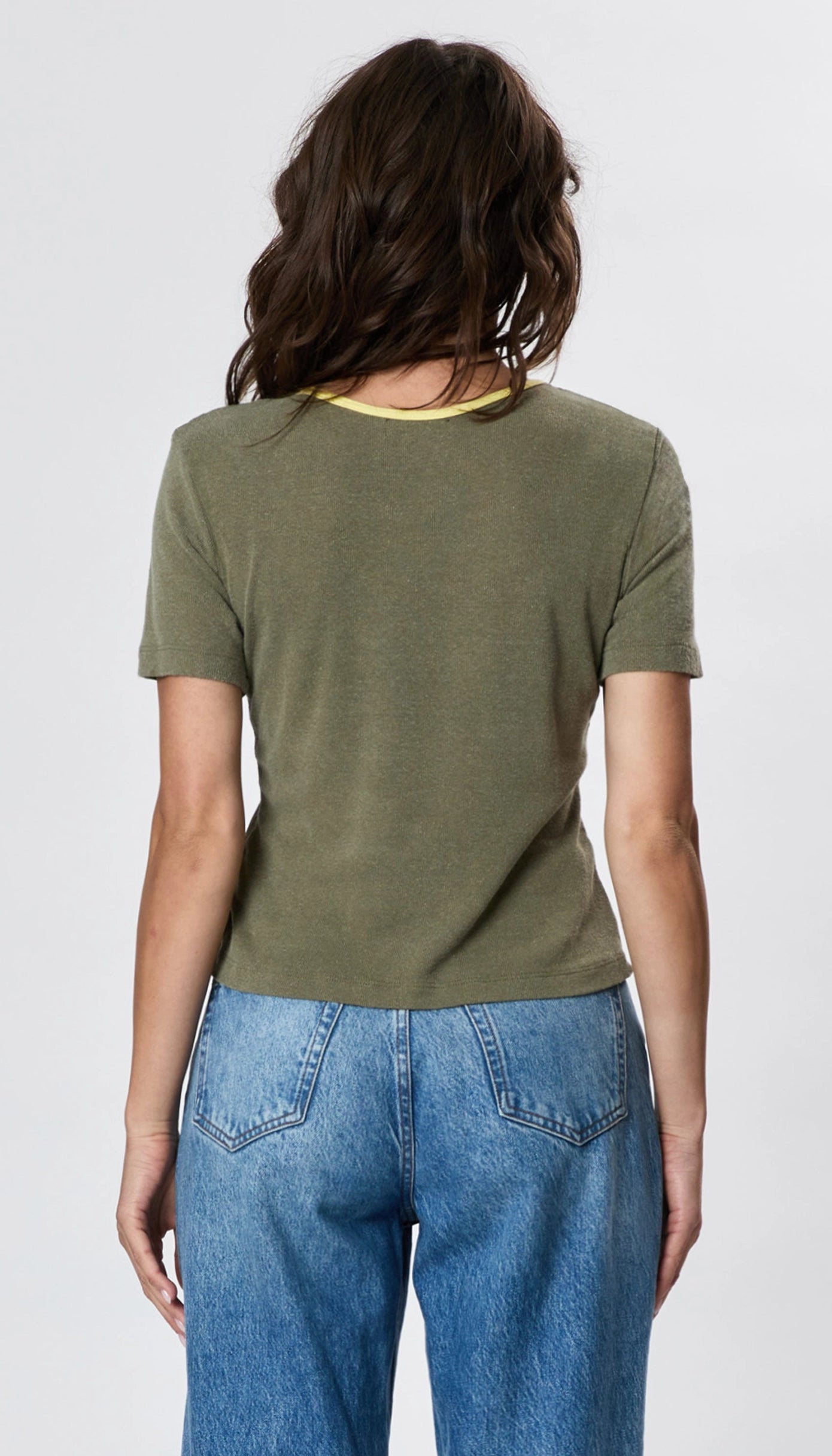 A woman with wavy brown hair is shown from the back, wearing Faire’s Stardust Hemp Top—an olive green short-sleeve tee with a yellow neckline—plus high-waisted blue jeans, radiating retro charm against a light background.