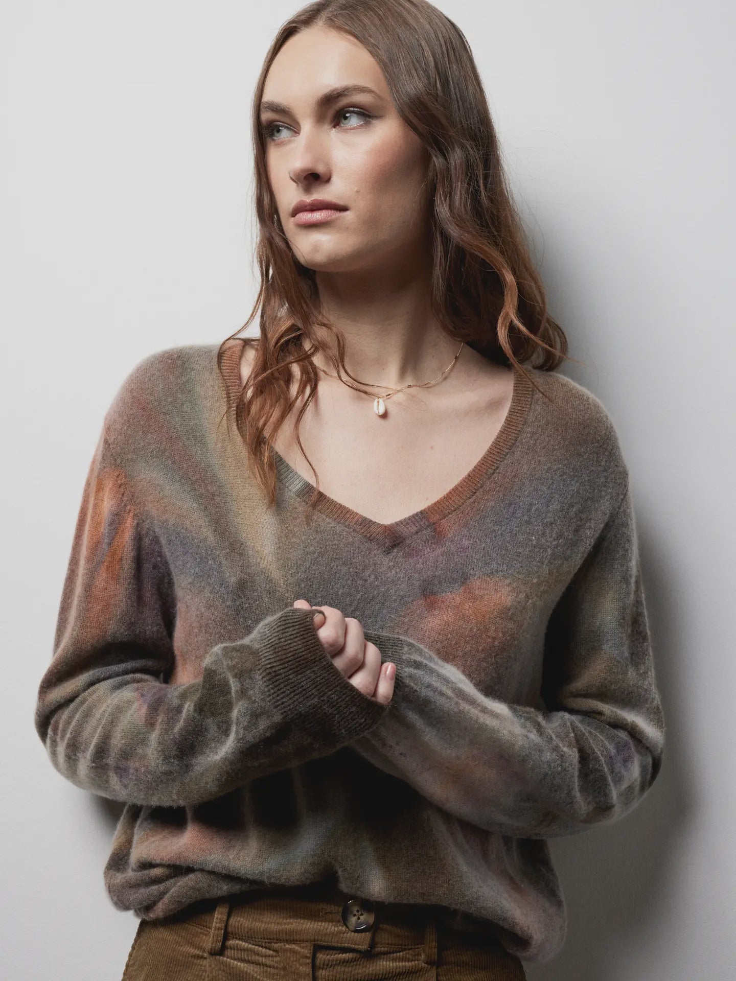 A woman with long, wavy brown hair wears the Riverside Tool & Dye Cashmere V-neck sweater, standing against a white wall and gazing to the side with a neutral expression.
