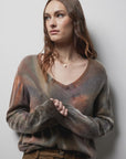 A woman with long, wavy brown hair wears the Riverside Tool & Dye Cashmere V-neck sweater, standing against a white wall and gazing to the side with a neutral expression.