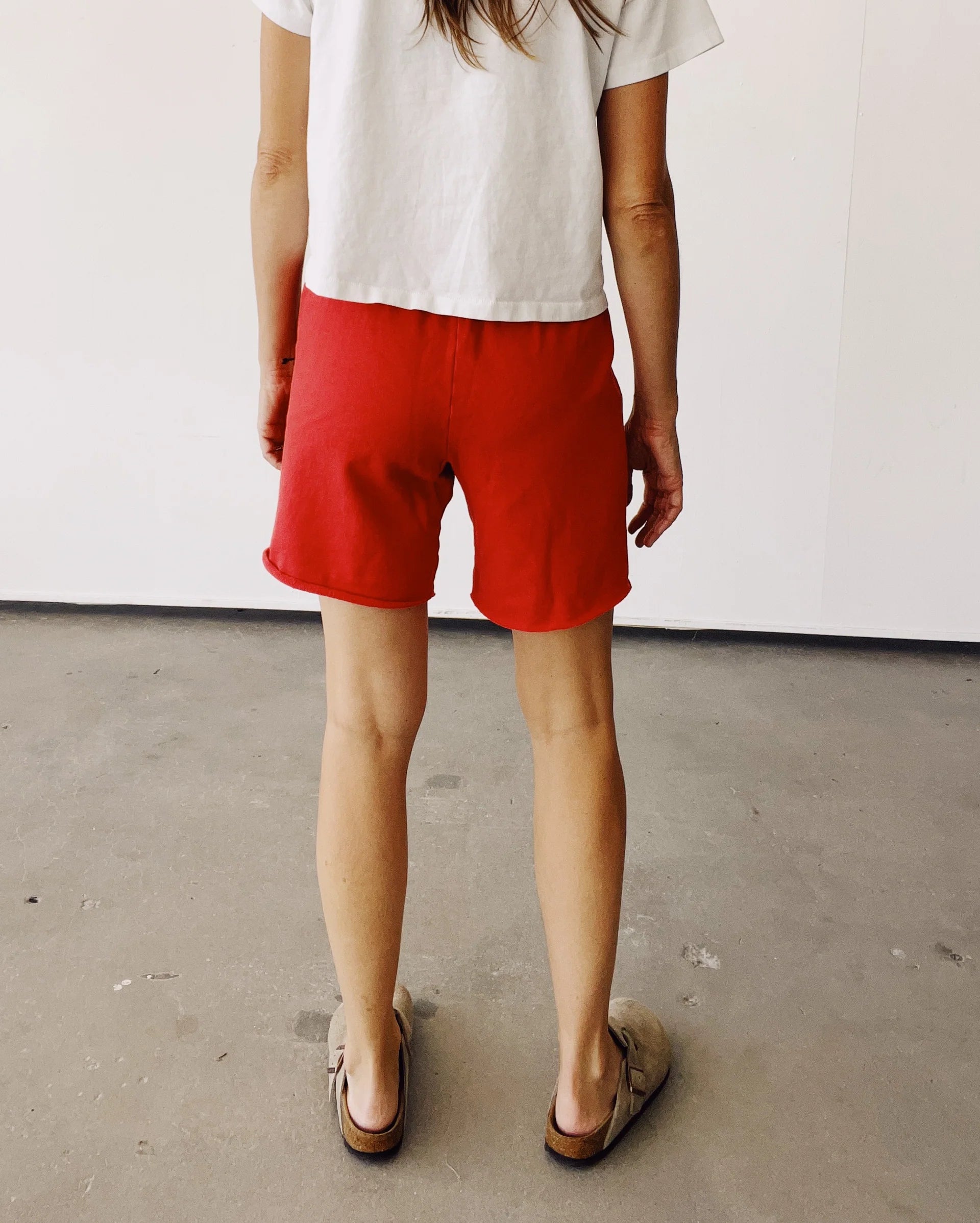 A person with long hair stands indoors on a concrete floor, wearing a white t-shirt and Hey Gang's The Sweat Shorts in red cotton with an elastic waistband, paired with tan slip-on sandals.