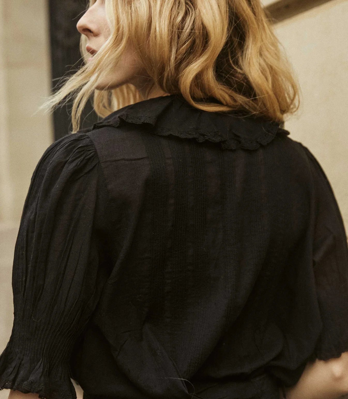 A person with wavy blonde hair wears the DOEN Henri Top, featuring an Edwardian-inspired ruffled collar, seen from behind and slightly in profile near a beige wall.