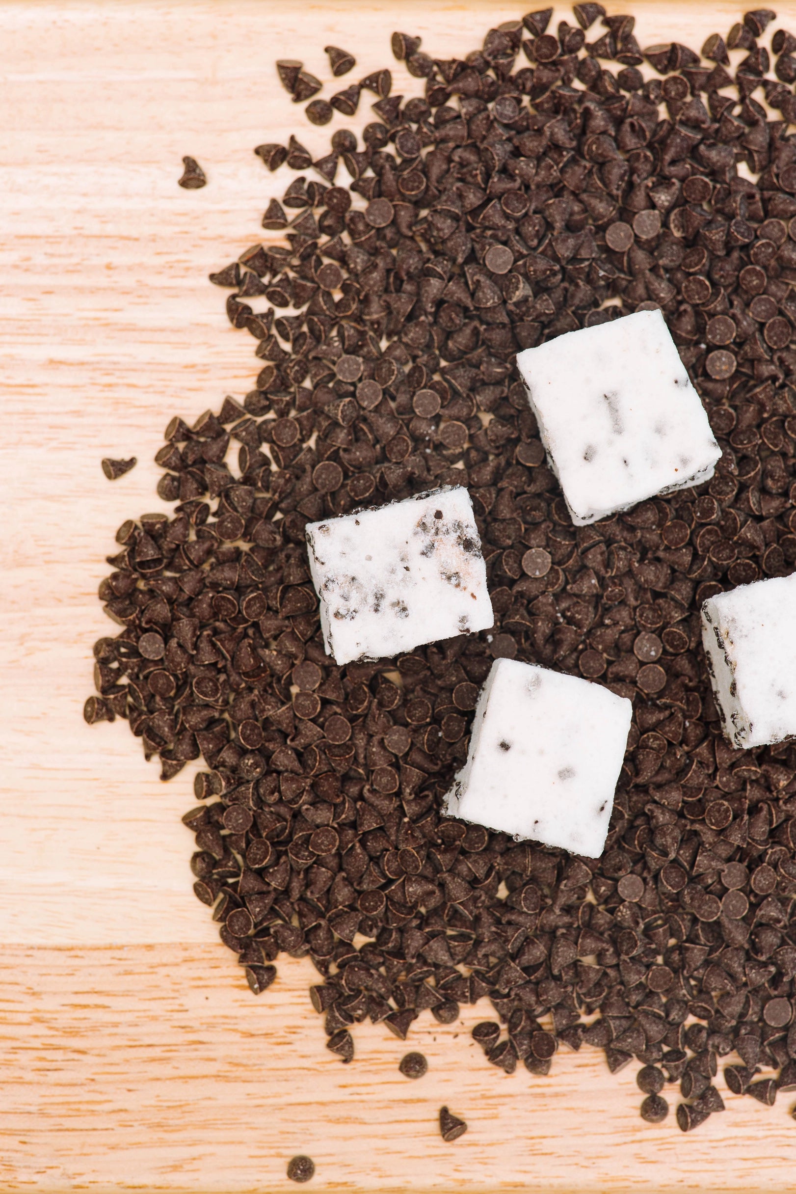 Four white, square Faire Choc Chip Marshmallows with chocolate specks sit on a bed of chocolate chips atop a light wooden surface.
