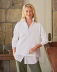 A person with blonde hair exudes a preppy vibe indoors, wearing a white collared Patrick Popover Henley by Frank & Eileen over a 100% cotton shirt and olive pants. They're leaning against a wooden chair, set against a stone wall with a table holding books and a vase in the background.