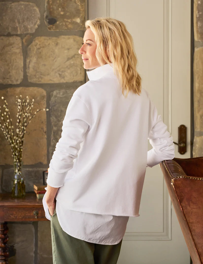 A person with blonde hair stands in a room with stone walls, wearing a Frank &amp; Eileen Patrick Popover Henley and green pants. A wooden table with a decorative vase and books sits beside them, while a leather chair partially adds to the preppy ambiance.