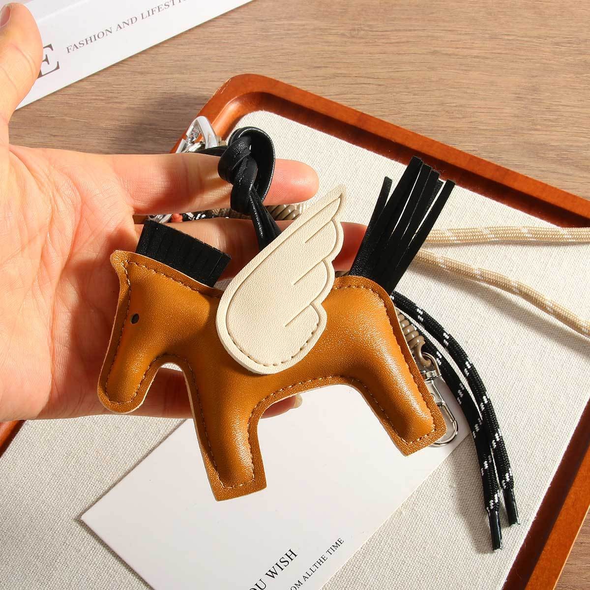 A hand holds the Faire Pegasus Keychain, a semi-handmade PU leather accessory shaped like a winged horse with black mane and tail, shown on a tray with cords and a white card.