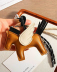 A hand holds the Faire Pegasus Keychain, a semi-handmade PU leather accessory shaped like a winged horse with black mane and tail, shown on a tray with cords and a white card.