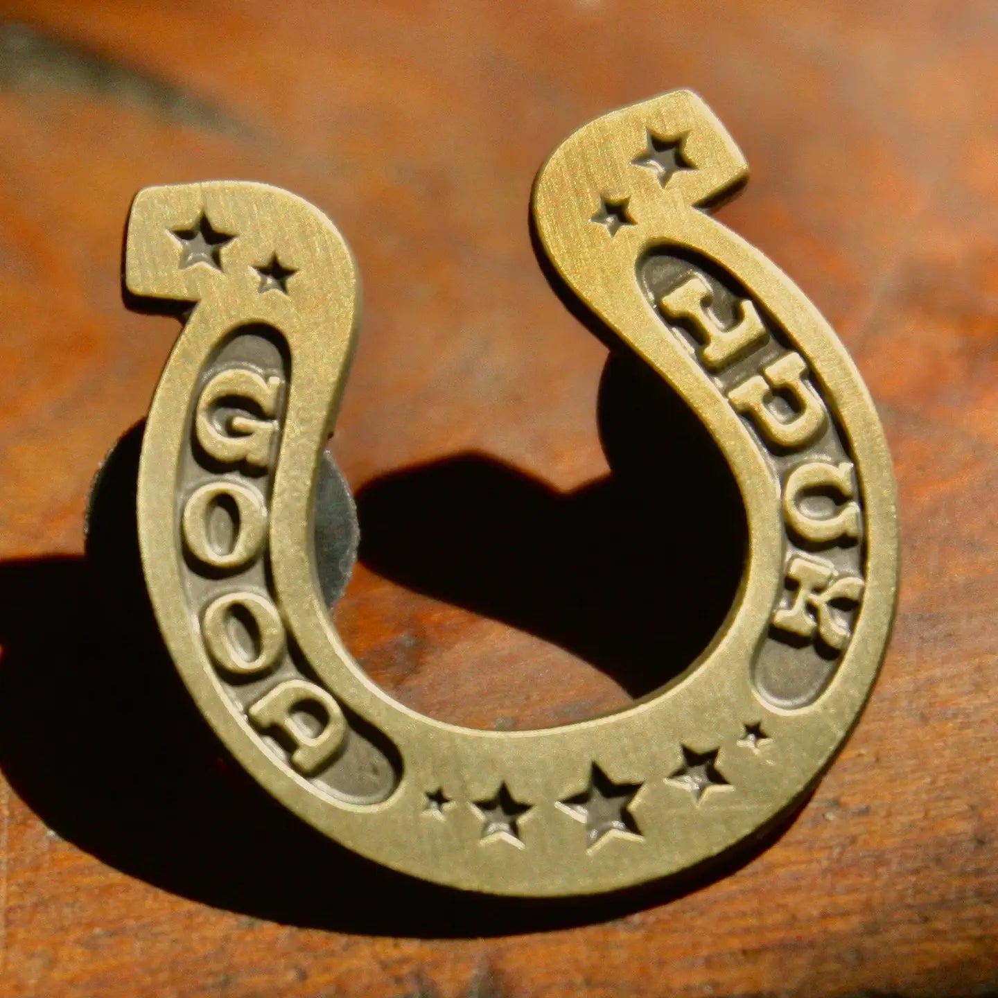 The Faire Good Luck Pin is a brass enamel pin engraved with “GOOD LUCK” and stars, shown resting on a wooden surface.