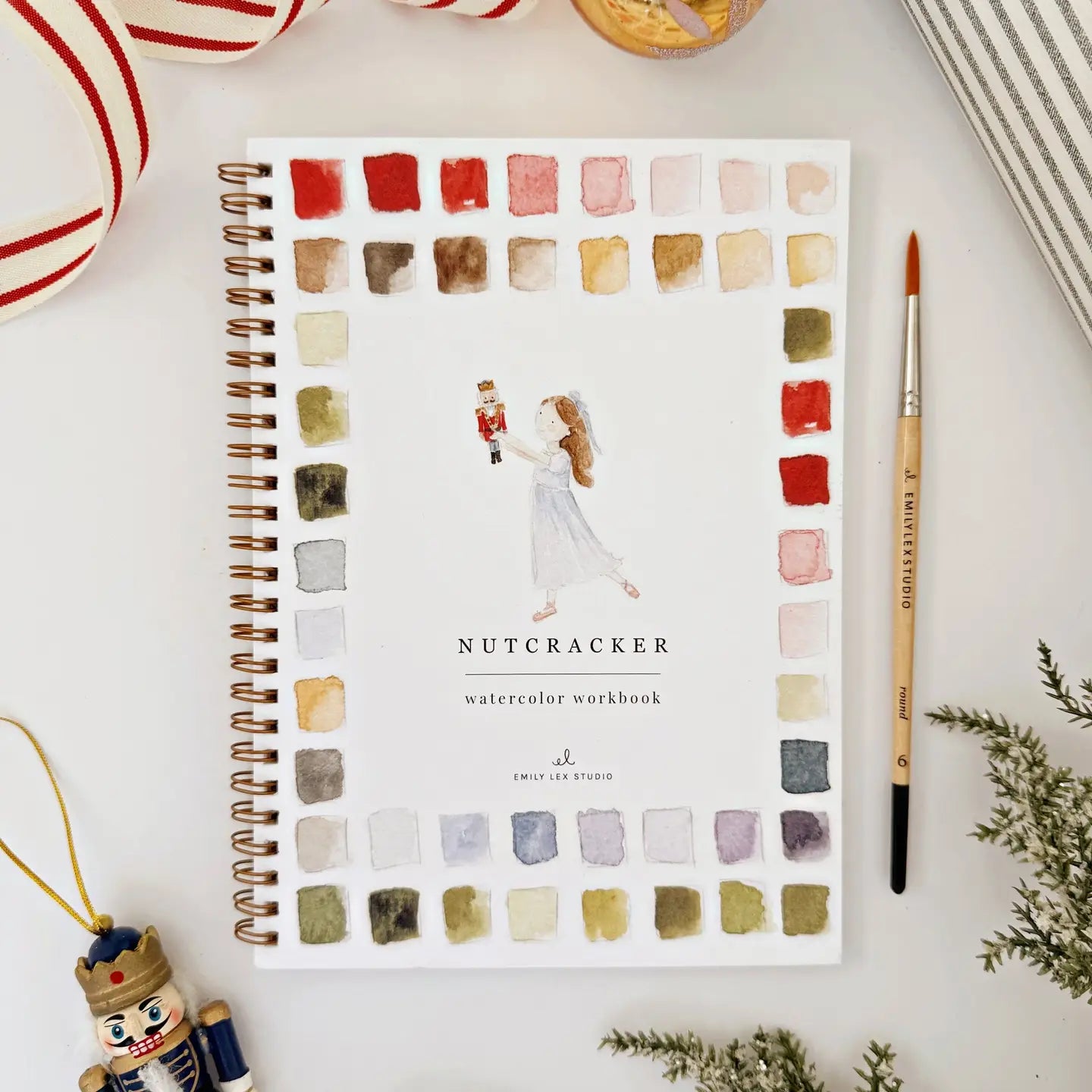 The Faire Watercolor Workbook, a spiral-bound beginner’s guide titled "Nutcracker," features a ballerina cover and paint swatches. Shown with a brush, nutcracker ornament, ribbon, and greenery for a festive touch.