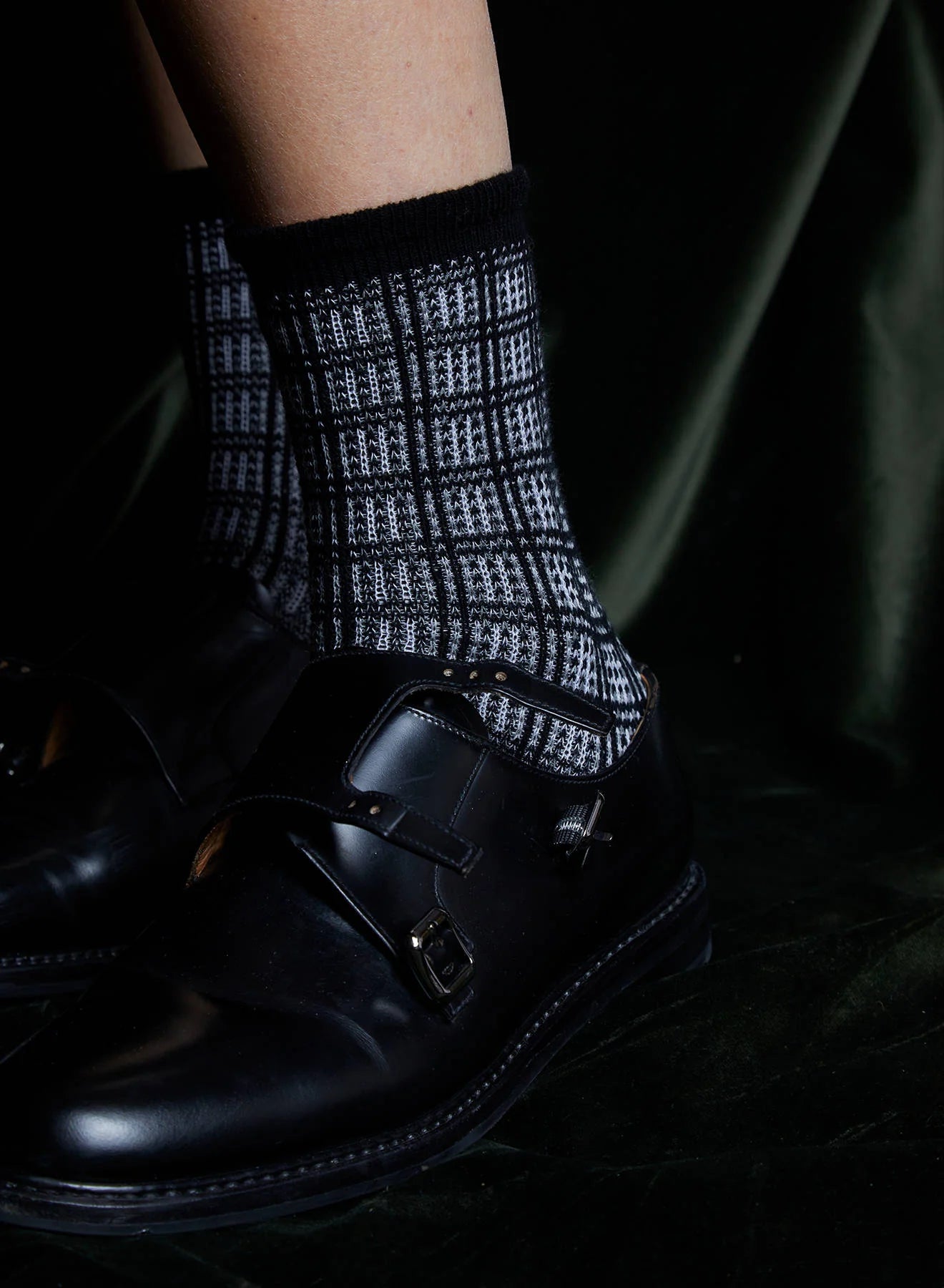 A close-up of a person’s lower leg wearing Maria La Rosa Iceland Socks—women’s mid-calf, black and white plaid merino wool—paired with black leather double monk strap dress shoes, set against a dark textured background.
