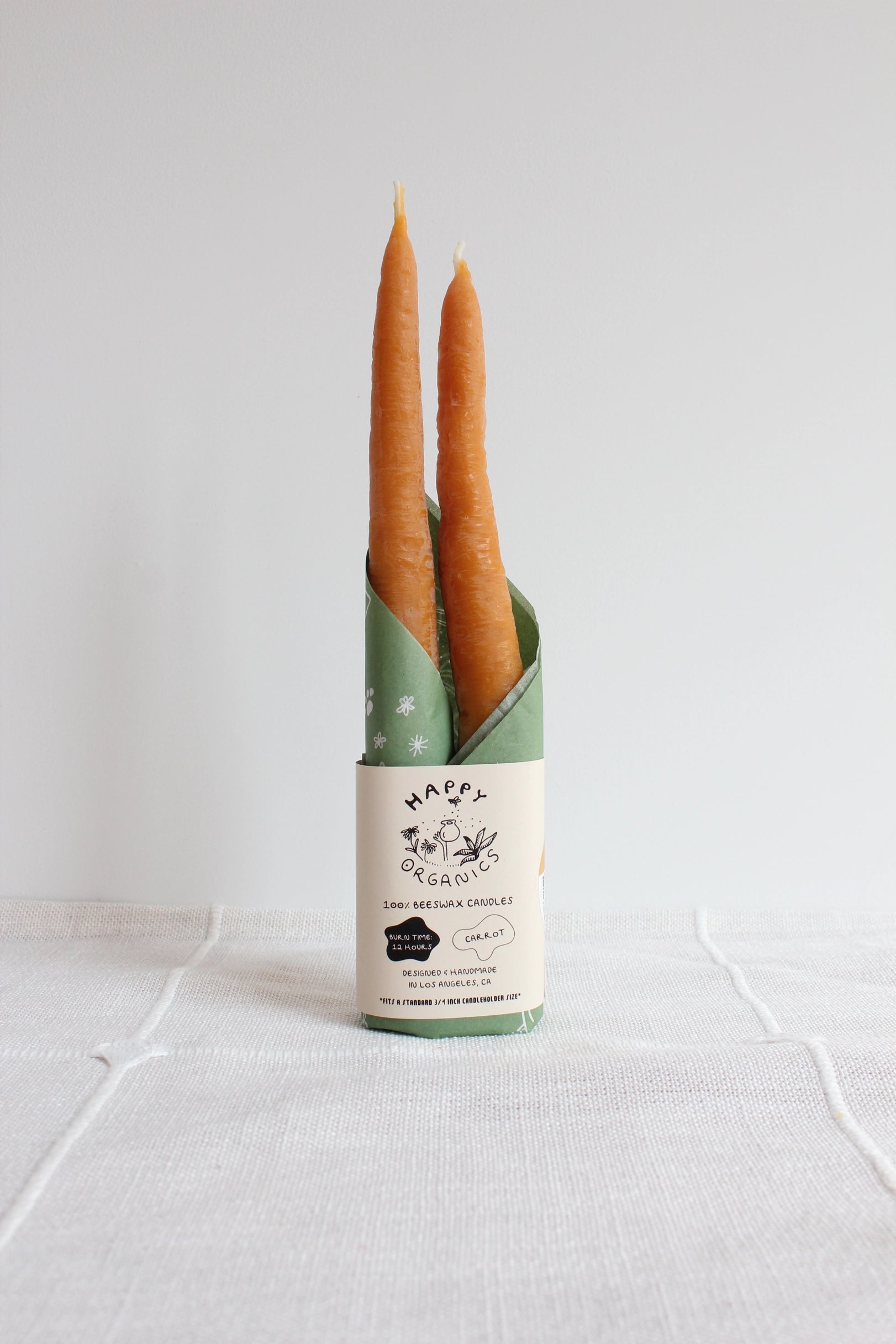 Two orange Carrot Beeswax Candles by Faire, made from beeswax, are wrapped in green paper and stand upright on a white textured surface against a plain white background.