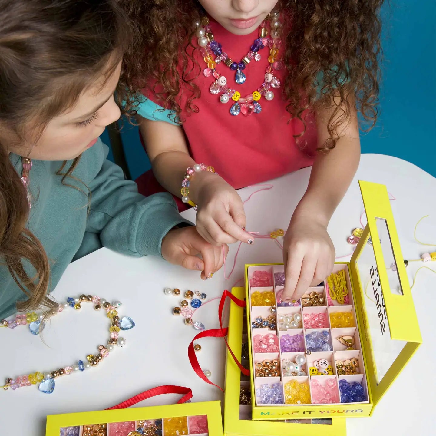 Two kids sit at a round table, crafting jewelry with colorful beads from the Make It Super DIY Bead Kit by Faire. They create and wear beaded necklaces and bracelets, engrossed in their vibrant, creative project.