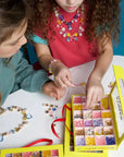 Two kids sit at a round table, crafting jewelry with colorful beads from the Make It Super DIY Bead Kit by Faire. They create and wear beaded necklaces and bracelets, engrossed in their vibrant, creative project.