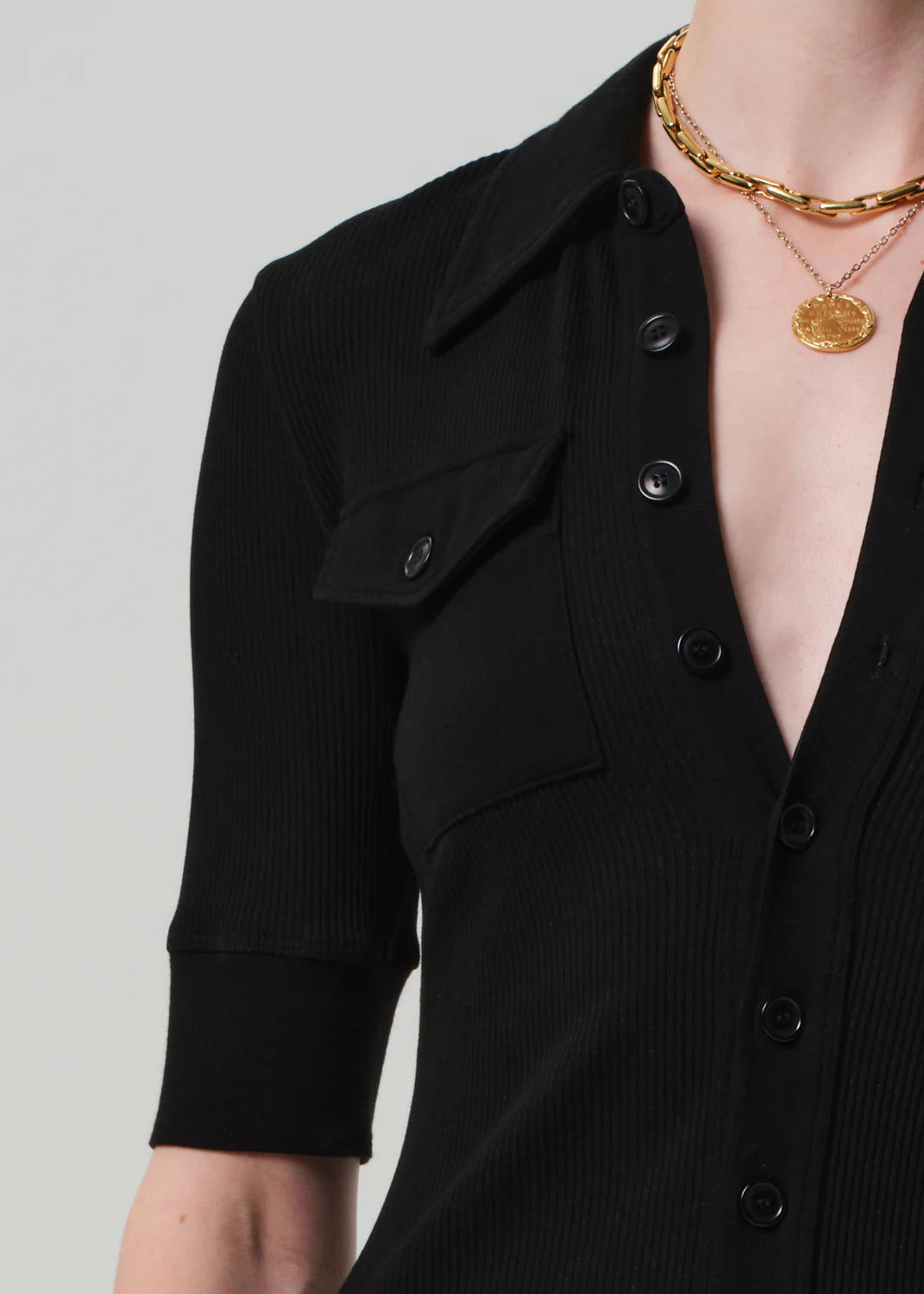 A person wears the Citizens Of Humanity/AGOLDE Amina Polo Dress—a black, ribbed cotton button-up with half sleeves and a chest pocket—paired with layered gold chain and coin pendant necklaces. The background is plain light gray.