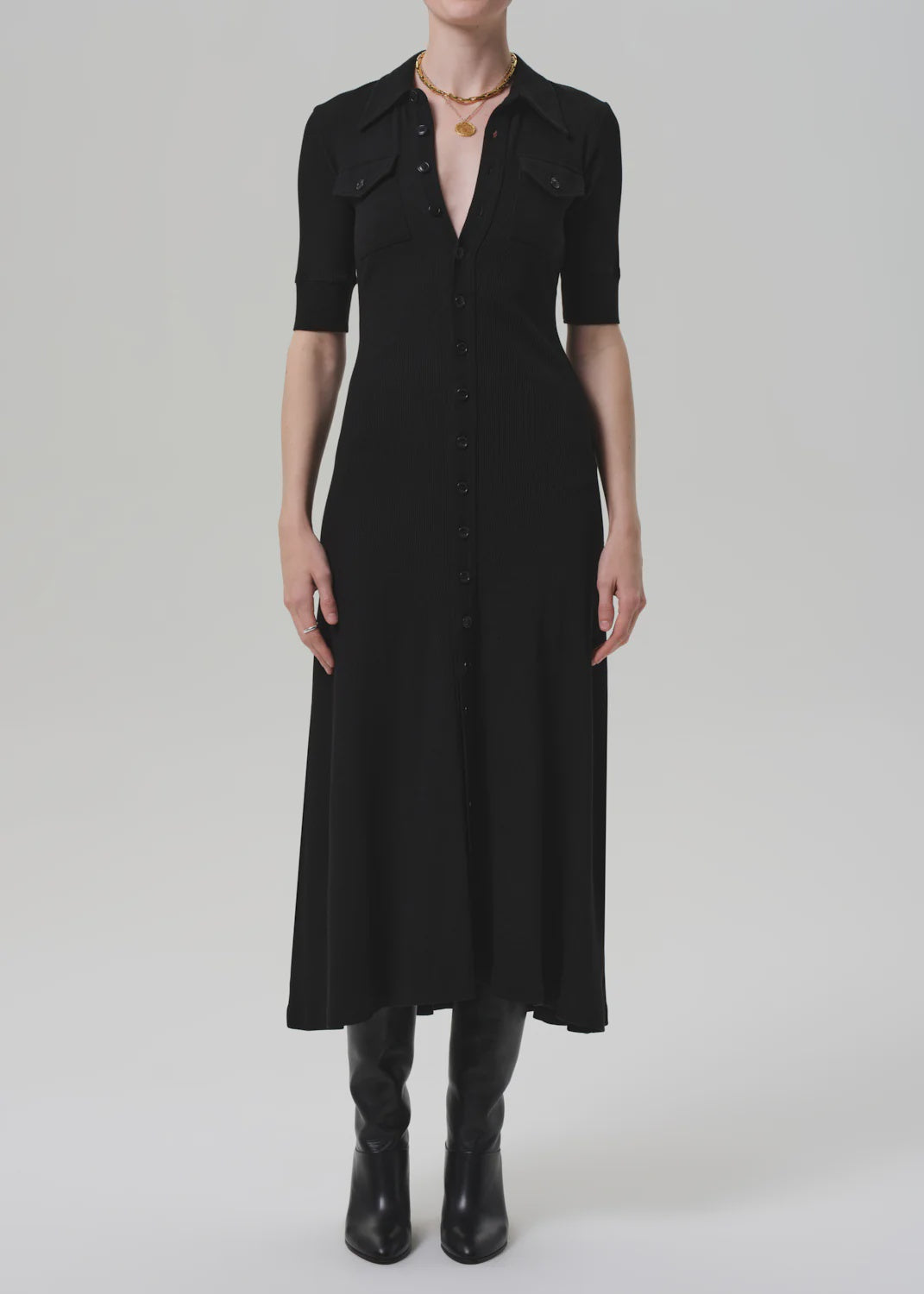 A woman in the Amina Polo Dress by Citizens Of Humanity/AGOLDE—fitted, black, with chest pockets—pairs it with a gold necklace and knee-high black boots against a plain light gray background.