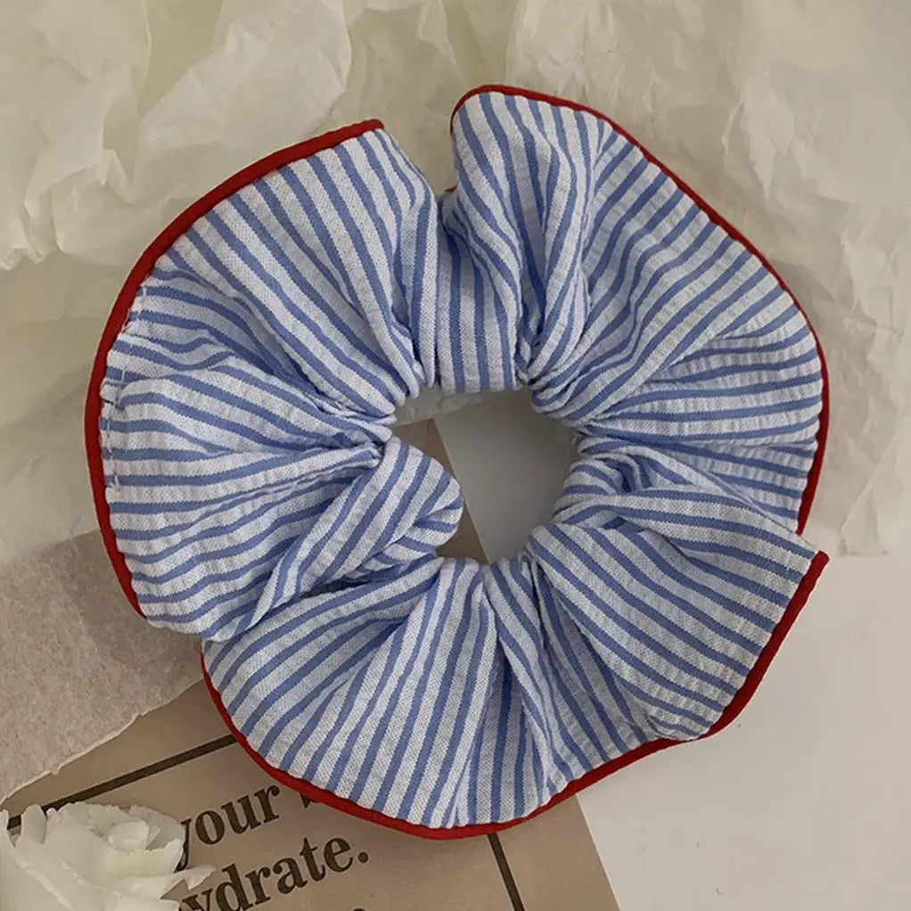 Faire Hair Scrunchies: A blue and white striped fabric scrunchie with red trim, perfect for fans of charming hair ties, is displayed on a light surface near paper and flowers.