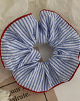 Faire Hair Scrunchies: A blue and white striped fabric scrunchie with red trim, perfect for fans of charming hair ties, is displayed on a light surface near paper and flowers.