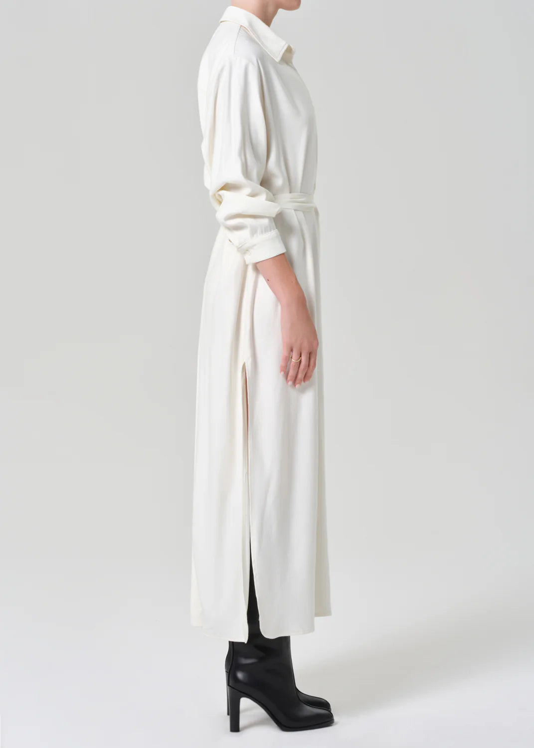 A person in profile wears the Citizens Of Humanity/AGOLDE Camillia Shirt Dress in Portofino Twill—a long, white belted dress with side slits—paired with black heeled boots against a plain light background.