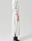 A person in profile wears the Citizens Of Humanity/AGOLDE Camillia Shirt Dress in Portofino Twill—a long, white belted dress with side slits—paired with black heeled boots against a plain light background.