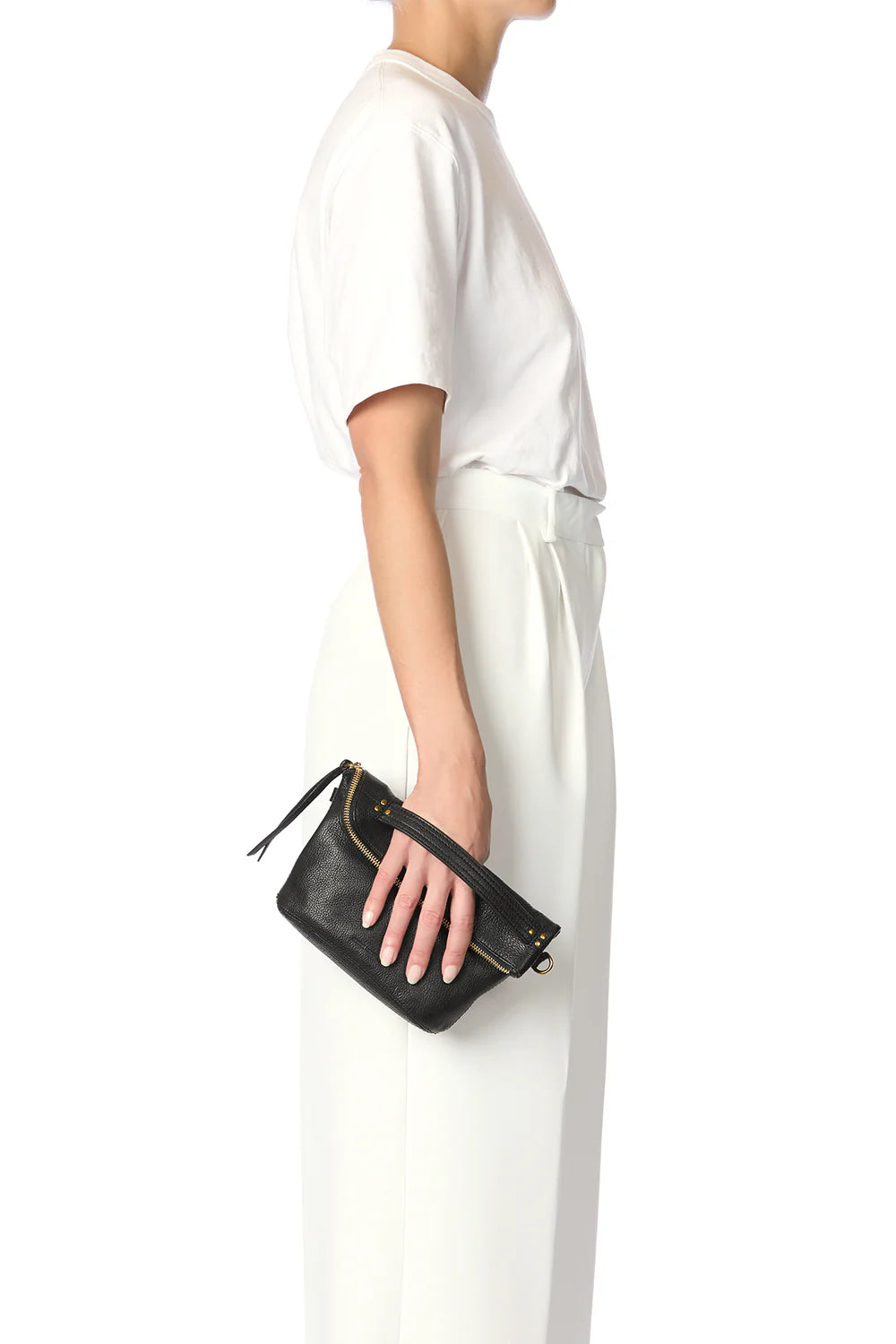 Wearing a white t-shirt and wide-legged high-waisted white pants, they hold the Jerome Dreyfuss Lucky Pochette—a small black goatskin handbag—against a plain white backdrop.