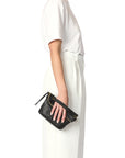 Wearing a white t-shirt and wide-legged high-waisted white pants, they hold the Jerome Dreyfuss Lucky Pochette—a small black goatskin handbag—against a plain white backdrop.