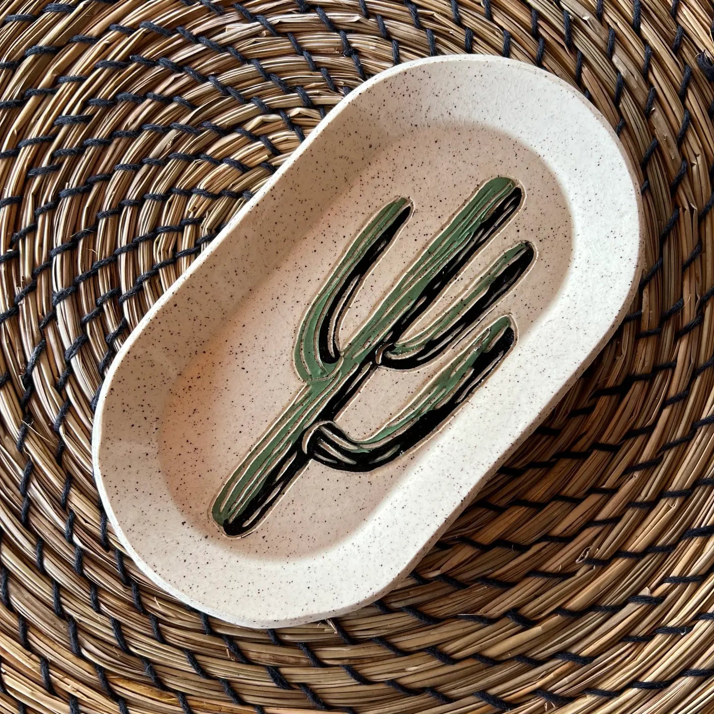 The Faire Saguaro Oval Trinket Dish features a painted green cactus design and rests on a round, spiral-patterned woven straw placemat, adding desert-inspired charm to any space.