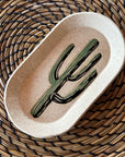 The Faire Saguaro Oval Trinket Dish features a painted green cactus design and rests on a round, spiral-patterned woven straw placemat, adding desert-inspired charm to any space.