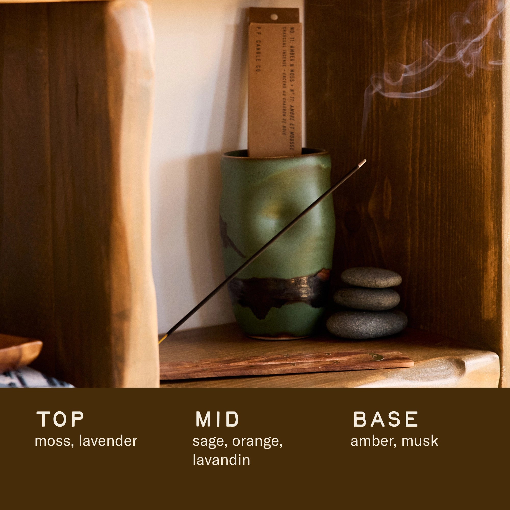 On a wooden shelf, Faire's PFC Incense Sticks burn in a green ceramic holder beside stacked stones. Scent notes: Top—moss, lavender; Mid—sage, orange, lavandin; Base—amber, musk.