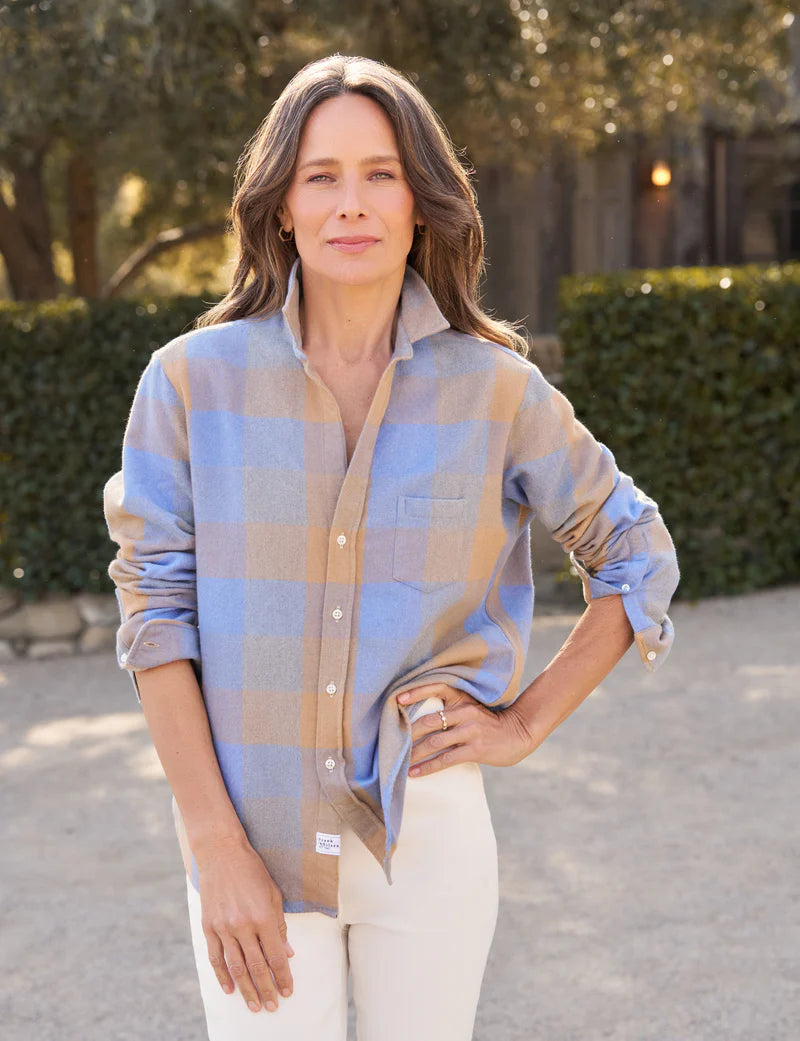 A woman with wavy brown hair stands outdoors in the Frank &amp; Eileen Eileen Button Up Shirt F25, featuring bust-flattering button placement and a relaxed fit, paired with white pants amid green bushes and trees.