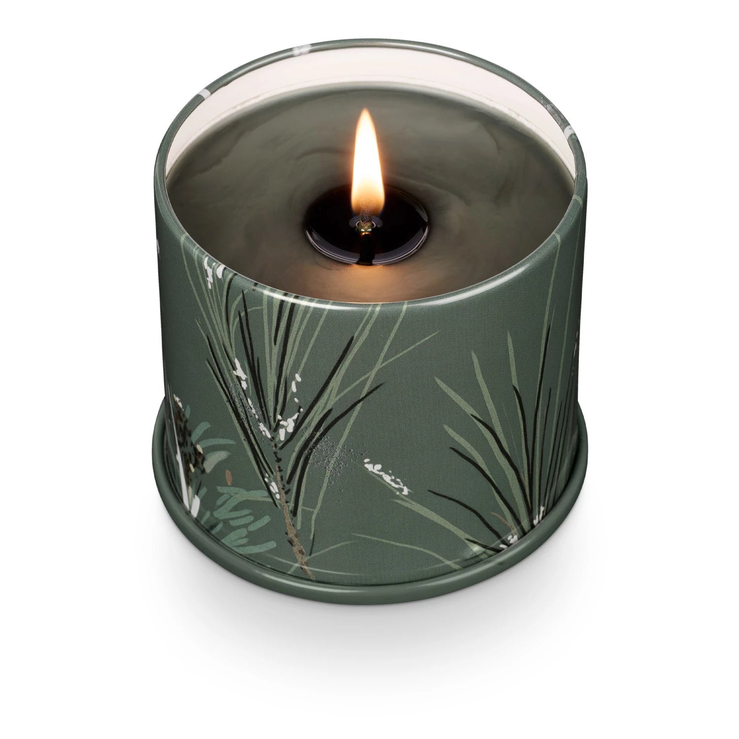 A lit Creative Co-op Vanity Tin Candle in a round, green holder adorned with botanical illustrations. The Balsam & Cedar scent fills the air, with the flame casting a cozy glow against the dark wax and creating a warm and inviting atmosphere.