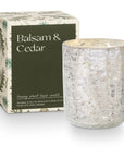 A Crackle Glass Candle Small by Creative Co-op is housed in a decorative, silver-textured holder beside a compact green and white box labeled "Balsam & Cedar," showcasing an elegant botanical design.