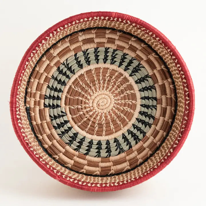 The Large Manuela Basket by Faire is a round, handwoven piece featuring tan, black, and pale green patterns, a wide red rim, and zigzag accents. Displayed on a white background.