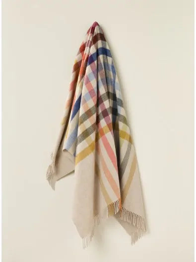 The Faire BM Merino Lamb Throw/Blanket, in beige with colorful red, blue, yellow, and green plaid stripes and fringed edges, is displayed neatly hanging on a light-colored wall.