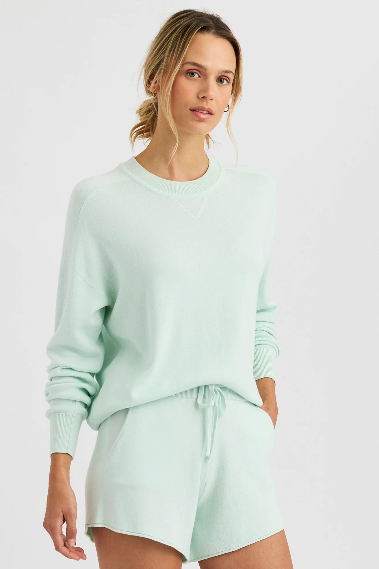 A woman stands against a white background, wearing the One Grey Day Warwick Pullover in light mint green with matching drawstring shorts; her hair is pulled back and her hands are in her pockets.