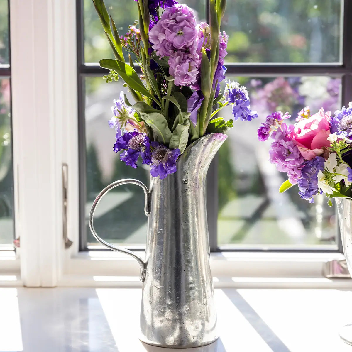 A Vintage Tall Tapered Pitcher by Faire, filled with purple and pink flowers, sits on a windowsill as sunlight streams in through the window behind it.