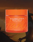 An orange jar labeled "Tallow Body Butter Moisturizer" by Faire, 4 fl oz (120 mL), made with grass-fed beef tallow and Sarasota honey, is shown before a silhouette of a person on horseback at sunset.