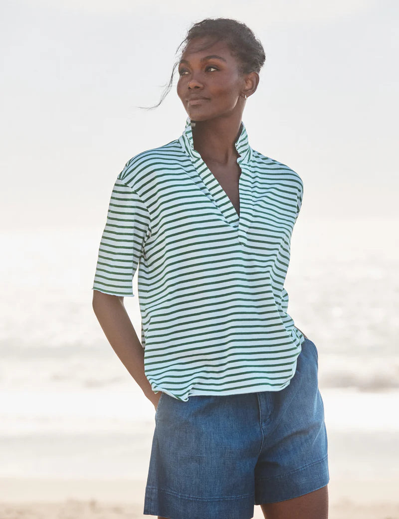 A person stands on a beach in a short-sleeved Patty Popover SS Henley by Frank & Eileen and blue shorts, hands in pockets, gazing to the side with the ocean blurred in the background.