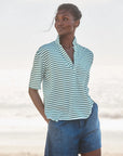 A person stands on a beach in a short-sleeved Patty Popover SS Henley by Frank & Eileen and blue shorts, hands in pockets, gazing to the side with the ocean blurred in the background.