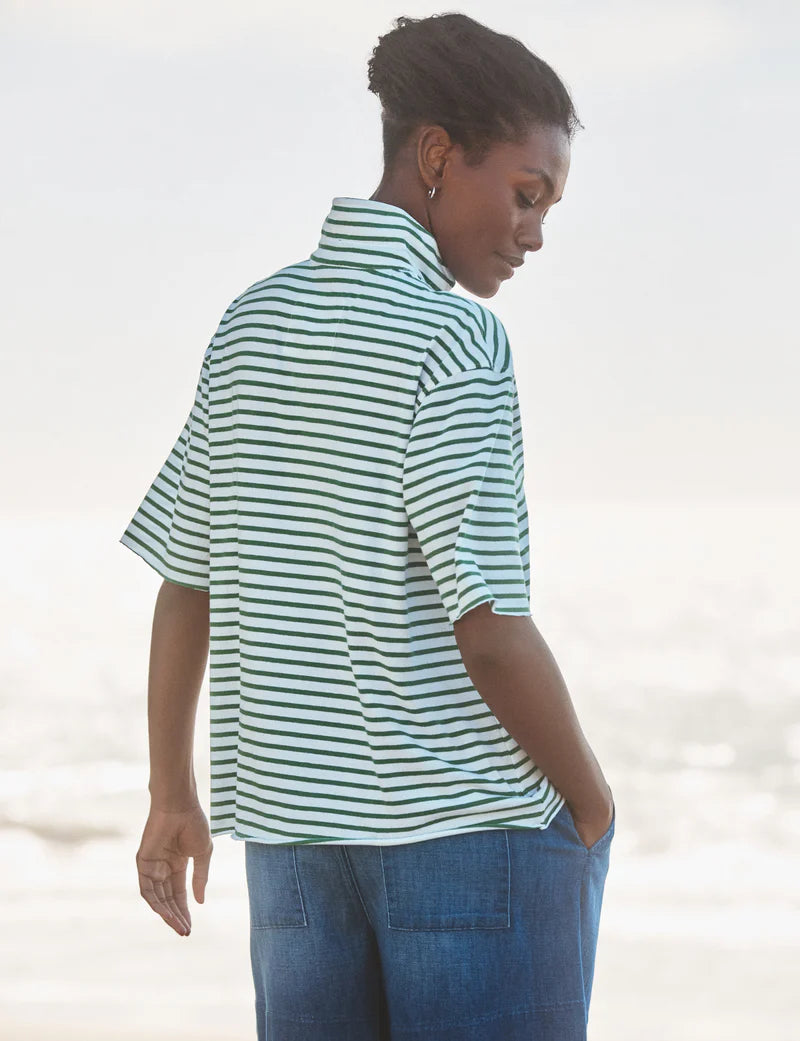 A woman stands outdoors with her back turned, wearing the Frank & Eileen Patty Popover SS Henley in mint-green and white stripes with blue pants. One hand is in her pocket as she looks over her shoulder toward the ground.