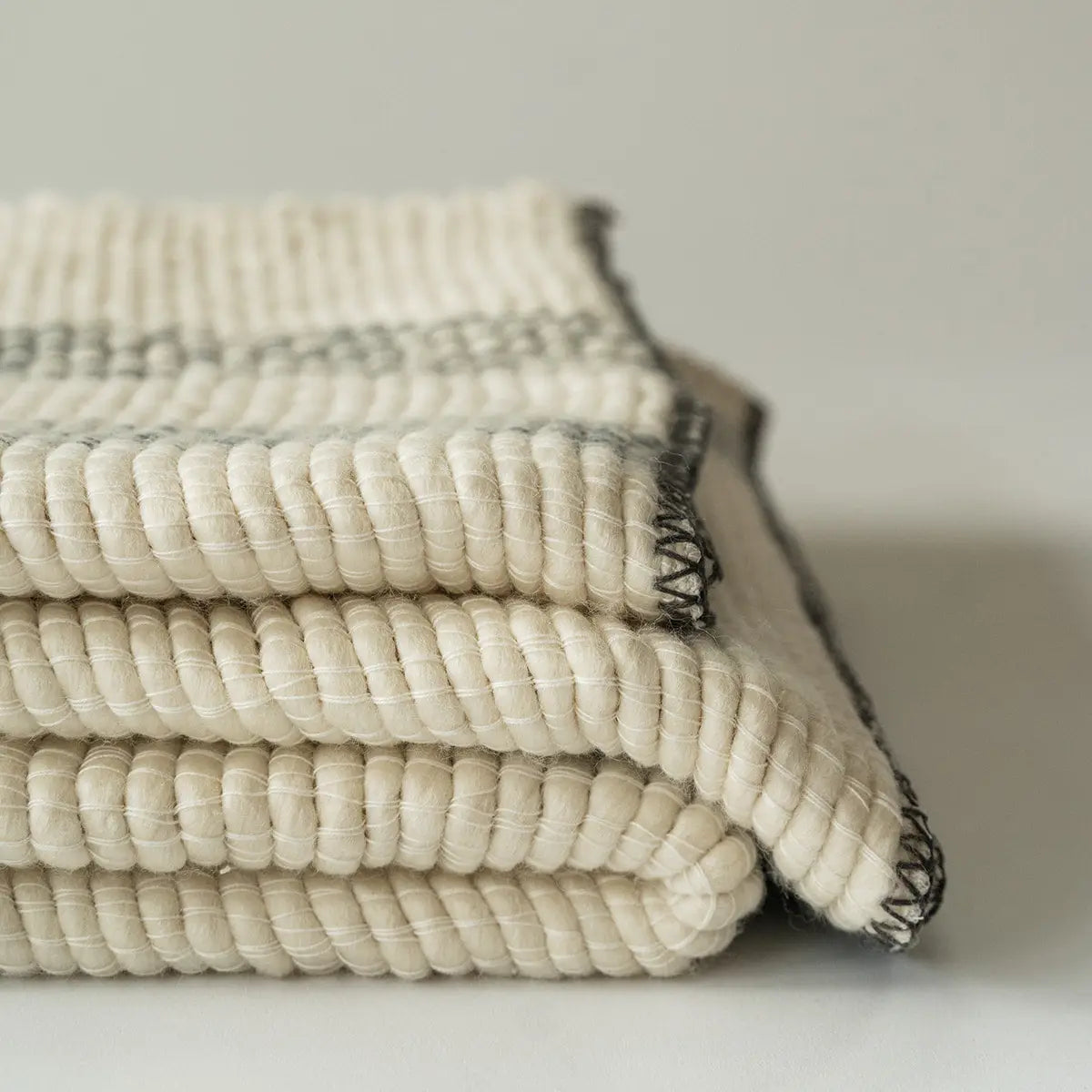 The Faire Cozy Stripe Throw, a thick cream-colored wool-blend with large visible stitches and dark edging, is neatly folded against a plain background.