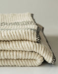 The Faire Cozy Stripe Throw, a thick cream-colored wool-blend with large visible stitches and dark edging, is neatly folded against a plain background.