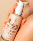 A hand holds a bottle of Faire Rahua Enchanted Island Body Glow Cream near a bare shoulder. This plant-based collagen booster offers anti-aging, youth-enhancing benefits.