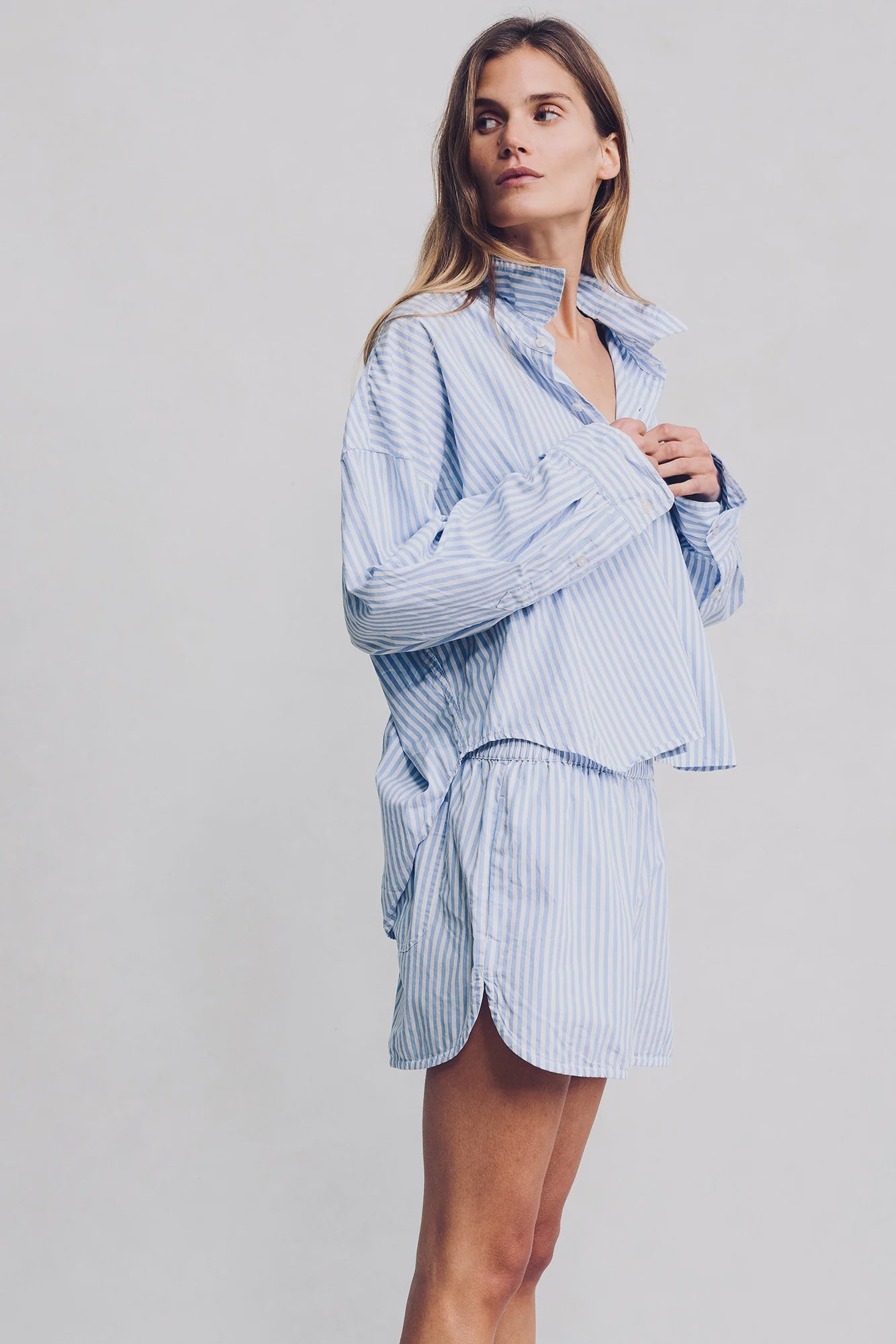 A woman wears Denimist PJ SHORTS—a loose-fitting, light blue and white striped cotton set—standing against a plain light background and looking slightly to the side.
