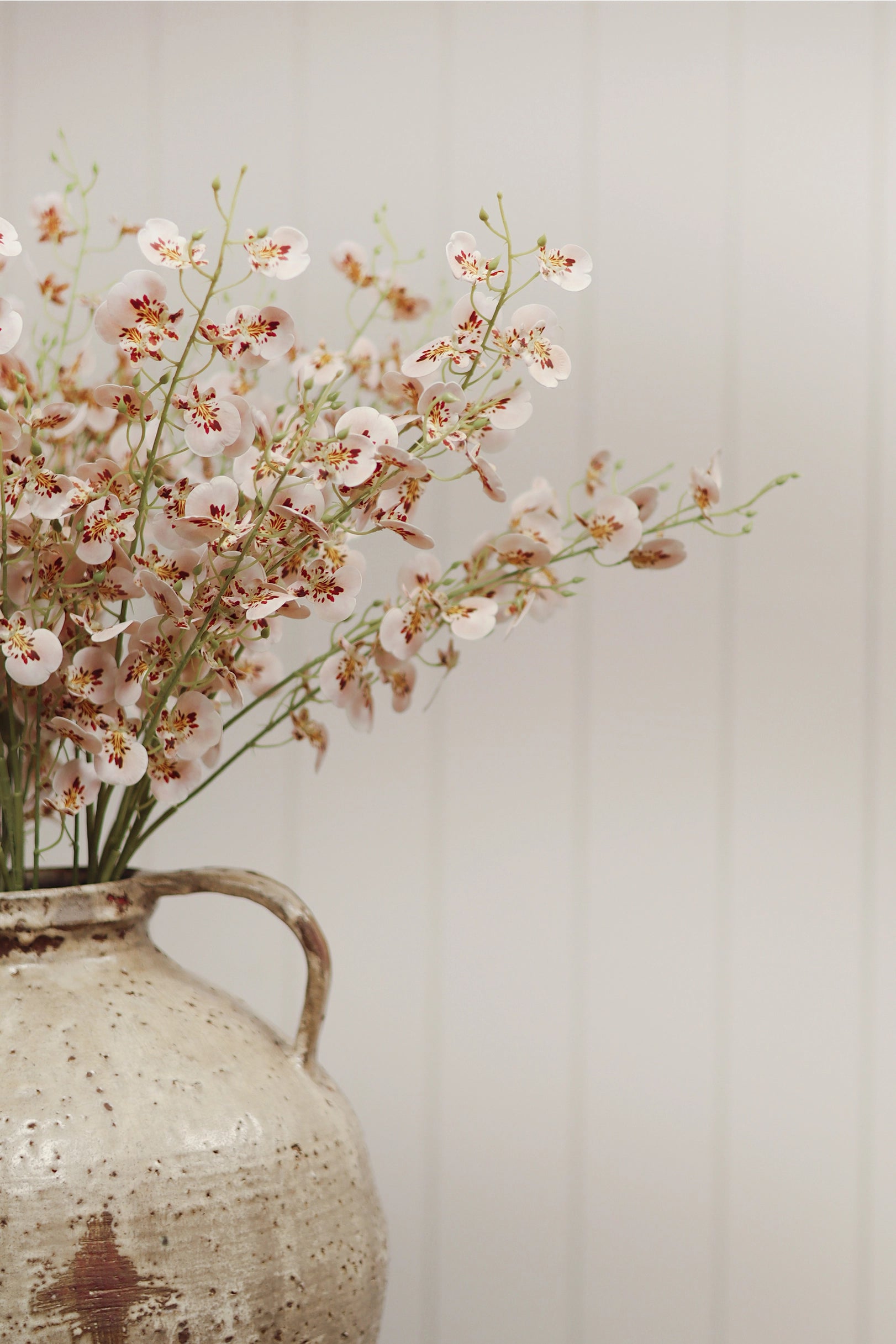The Faire Wild Orchid Spray features delicate pink and white faux flowers with slender green stems, arranged in a rustic ceramic vase and set against a light, vertically paneled background for an airy home decor accent.